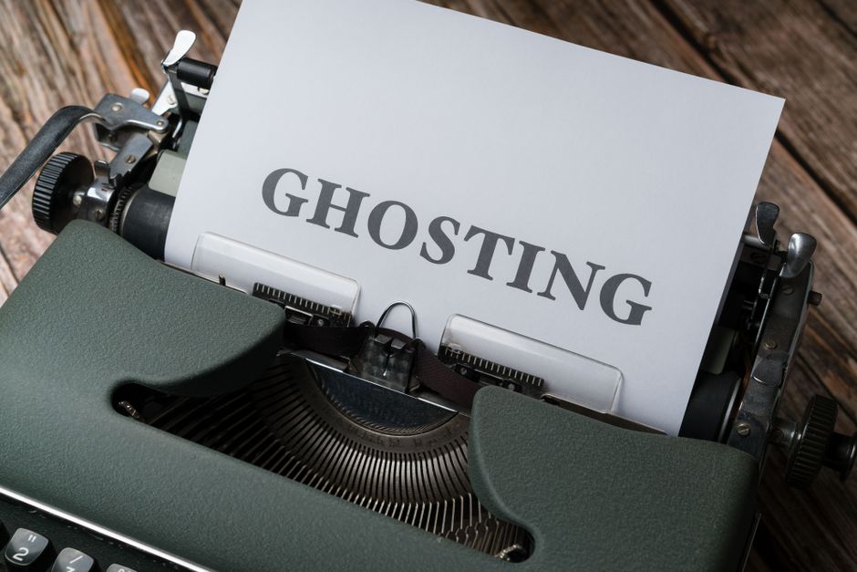 A vintage green typewriter positioned on a wooden surface, with a sheet of white paper inserted into the roller that has the word 'GHOSTING' printed on it in bold, black letters. The typewriter's keys, carriage, and side knobs are visible, with some metallic components reflecting light. The setting suggests an environment related to writing or documentation, which may relate to the process of packing, moving, or home relocation logistics. Harringay Man and Van, a professional removals company, may use such images to illustrate aspects of packing or moving services, including detailed handling and careful transport of items during house removals.