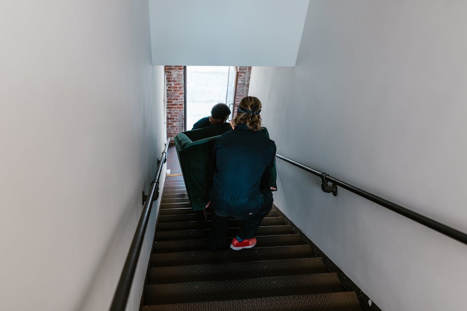 Two individuals engaged in a home relocation process are visible on a narrow indoor staircase with brown carpeted steps and white walls. The person in the foreground has long, curly hair covered with a headband, and is dressed in dark clothing with red footwear, crouching while holding the edge of a large, dark green upholstered armchair. Behind, another person is seen wearing a dark top, assisting with the furniture move. The staircase has black metal handrails on both sides, and at the top, natural light filters through a window that reveals exposed brickwork, indicating the building's interior walls. The scene captures the loading process, where furniture such as an armchair is being carefully carried down the stairs, supporting the overall home relocation or furniture transport effort by Harringay Man and Van, as part of their removals services for tight stair moves in Harringay.