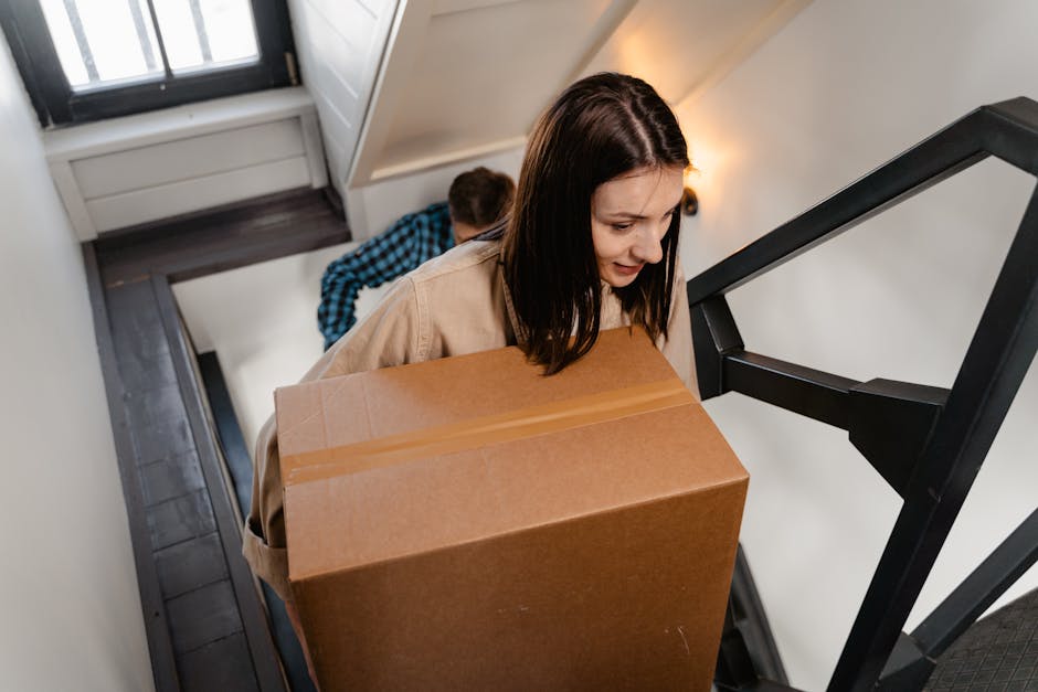 A woman with shoulder-length brown hair, wearing a beige coat, is carrying a large cardboard box up a narrow staircase inside a house. Behind her, a man dressed in a blue plaid shirt is also ascending, holding another box. The staircase has a black metal railing and wooden steps, with white walls on either side. Natural light from a window nearby illuminates the scene. Various packing materials, such as plastic wrap and cardboard boxes, are visible, indicating an ongoing packing and home relocation process. The image captures the careful handling of furniture and boxes during the loading process, supported by Harringay Man and Van's local removals service at harringaymanandvan.co.uk, focusing on tight stair moves in Harringay.