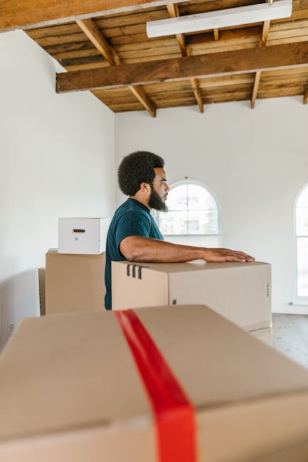 A man with curly hair and glasses, wearing a dark blue t-shirt, stands inside a bright, empty room with white walls and a wooden ceiling framework. He is leaning with his forearms on a large cardboard box, preparing for a furniture transport or home relocation process. Several other cardboard boxes, some with white labels, are stacked nearby at different heights, indicating packing and moving preparations. The room has two arched windows allowing natural light to illuminate the space, and the floor appears to be light wood or laminate. This scene depicts the loading process involved in house removals, supported by [COMPANY_NAME], highlighting the careful handling and organization typical in professional removals and packing services.