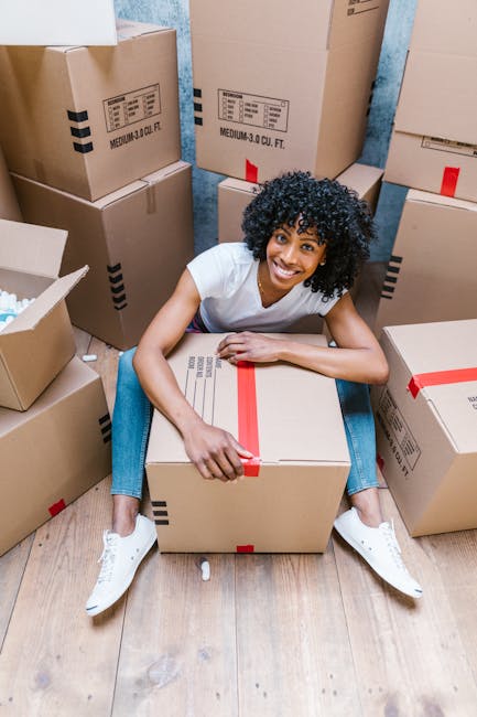 A man with curly hair and glasses, wearing a dark blue t-shirt, stands inside a bright, empty room with white walls and a wooden ceiling framework. He is leaning with his forearms on a large cardboard box, preparing for a furniture transport or home relocation process. Several other cardboard boxes, some with white labels, are stacked nearby at different heights, indicating packing and moving preparations. The room has two arched windows allowing natural light to illuminate the space, and the floor appears to be light wood or laminate. This scene depicts the loading process involved in house removals, supported by [COMPANY_NAME], highlighting the careful handling and organization typical in professional removals and packing services.