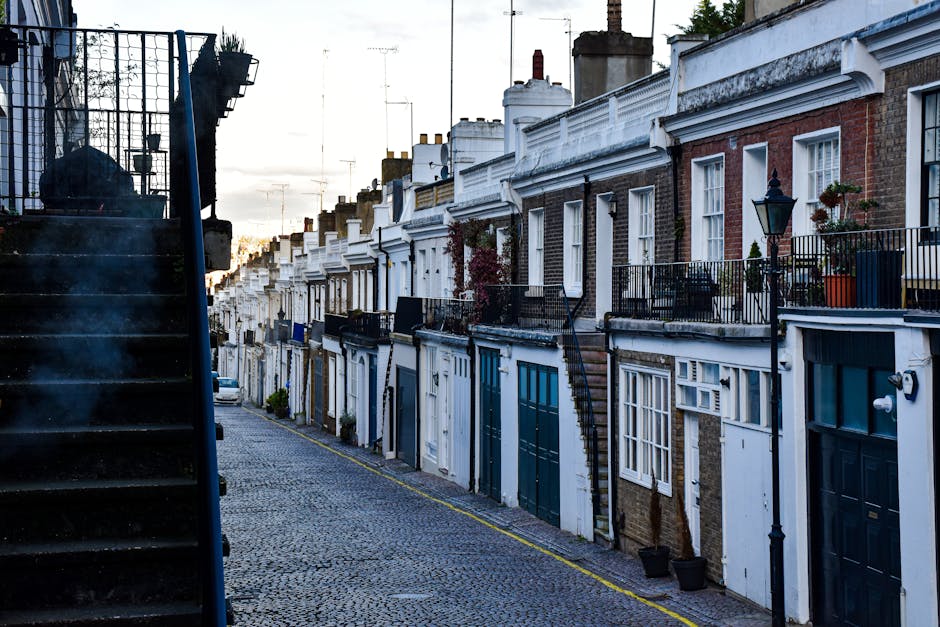 A narrow residential street with a steep incline featuring tightly packed terraced houses with brick and white facades, each with small front gardens or balconies decorated with potted plants and outdoor lighting. The street is paved with cobblestone and lined with a yellow guide line along the kerb. On the left side, a black metal staircase with visible steps and a railing leads to an upper level of a nearby building, with a large black bag resting on the staircase landing. On the right side, there are black wrought-iron street lamps attached to the front walls of the houses, which have multiple white-framed windows and doors painted in shades of blue and white. The scene appears to be illuminated by natural daylight, possibly during early evening as the sky shows faint light. This setting reflects typical urban housing, suitable for house removals and furniture transport, and highlights the logistical considerations of home relocation in narrow streets, aligning with services offered by Harringay Man and Van.