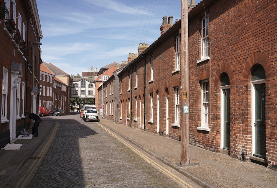 A quiet residential street featuring a row of traditional red-brick terraced houses with white-framed windows and front doors, set on a cobbled pavement under a clear blue sky. On the left side, a person is seen handling cardboard boxes, possibly packing or preparing items for a home relocation. The street is narrow with double yellow lines along the edge of the pavement, and a small white van is parked further down the street, positioned near the entrance to one of the houses. A wooden utility pole stands on the right side, with a small sign attached, and in the background, a modern, multi-story building with large windows spans across the street, connected by an overhead footbridge. The scene is well-lit, showing typical urban surroundings suited for furniture transport and household removals, aligning with the services offered by Harringay Man and Van.