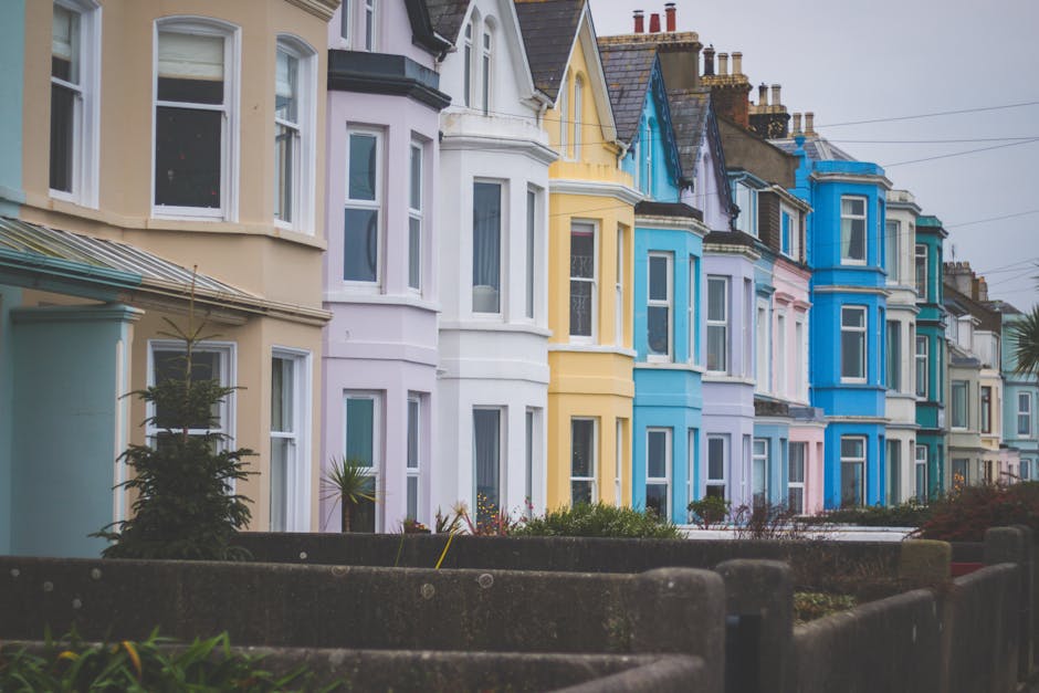 Image showing a row of Victorian terraced houses in Harringay, painted in pastel shades including light blue, yellow, white, pink, and purple, located in a residential area with small front gardens separated by concrete or stone boundary walls. The street appears to be overcast, with diffused natural light and overhead power lines visible. In the foreground, there are low concrete planters with some small shrubs and plants, and the pavement extends along the front of the houses. As part of a house removal or home relocation process, this scene could depict the exterior environment where furniture transport and loading activities might take place, with Harringay Man and Van providing professional moving services to facilitate packing, loading, and transport of belongings from these Victorian homes.