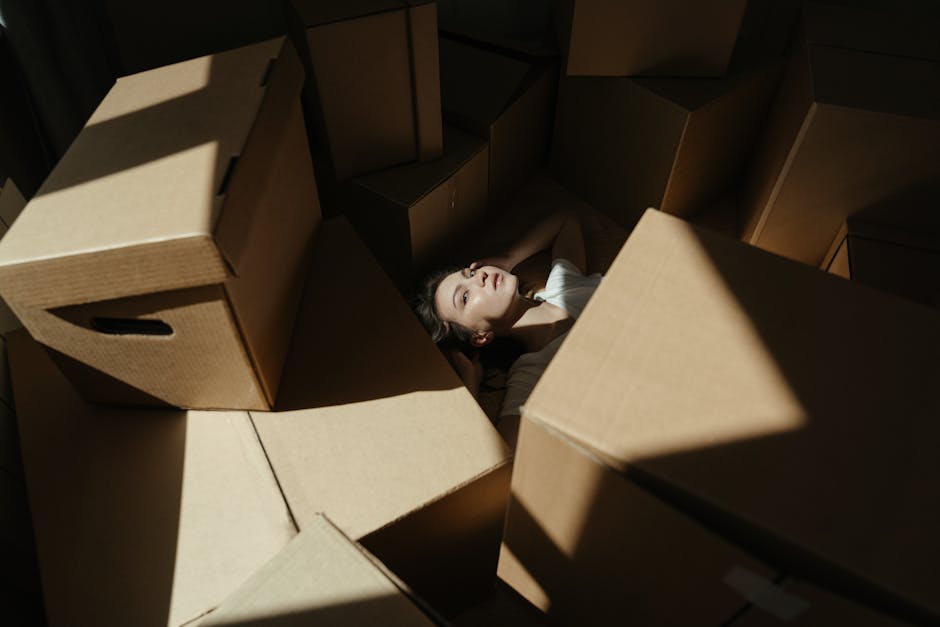 A woman with dark hair and a white top lying inside a large pile of flattened cardboard boxes and packing materials, surrounded by various cardboard boxes of different sizes, with some upright and others lying flat. She appears relaxed with her eyes closed and arms resting behind her head. The scene is set in a well-lit environment, possibly inside a property during a home relocation, with natural light casting shadows on the boxes and the woman's face. The image captures the packing and moving process, showing the careful handling of household items, as might be coordinated by a professional removals company such as Harringay Man and Van, in preparation for furniture transport and home relocation. The setting emphasizes the logistics involved in loading and organizing packed boxes for a move, with a focus on the packaging materials, the spatial arrangement of the boxes, and the environment where packing activities are taking place.