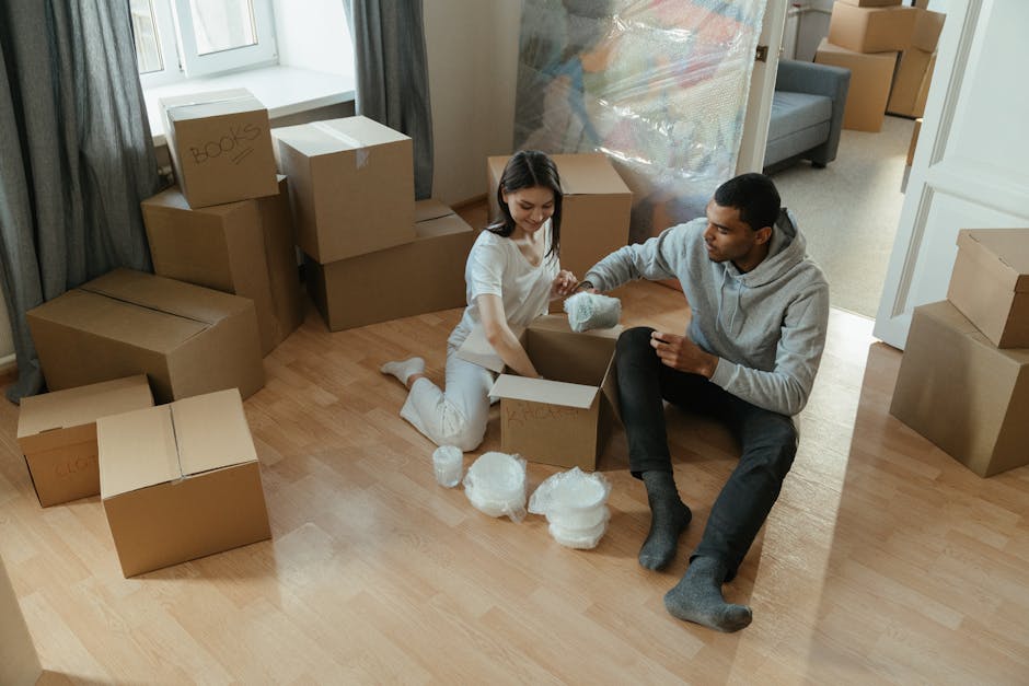 A young woman and a young man are sitting on a light wooden floor inside a home, surrounded by numerous cardboard boxes used for packing during a house relocation. The woman is opening one of the boxes, which contains bubble wrap, and she is holding a piece of bubble wrap in her hand. The man is assisting her by holding a small item wrapped in bubble wrap, possibly for moving or storage. Several other packed boxes, some labeled with black marker, are stacked against the wall near a window with grey curtains, indicating an ongoing packing process. In the background, part of the living room is visible with a grey sofa, a plastic wrapping sheet hanging near the doorway, and scattered packing materials on the floor. The scene depicts a typical packing and loading process in preparation for furniture transport as part of a home or flat move, facilitated by Harringay Man and Van, a specialist in removals and relocation services.