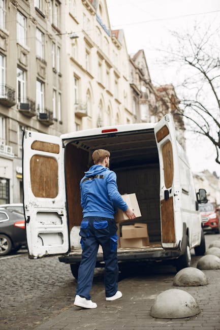 A man dressed in a blue jacket, dark blue trousers, and white sneakers is seen loading cardboard boxes into the open rear of a white cargo van parked on a cobbled street in Harringay. The van's rear doors are wide open, revealing the interior lined with wooden panels, and several packed boxes are being placed inside as part of the home relocation process. The street is lined with multi-storey residential buildings with light-colored facades and bay windows, and there are parked cars along the sides of the road. Bare trees and overcast sky are visible in the background, indicating a typical urban environment. The scene captures the logistics of furniture transport and packing during a moving service, with visible equipment including the boxes and the van designed for efficient loading and transport. Harringay Man and Van's removal service is implied as they handle the loading process in this typical urban moving scenario.