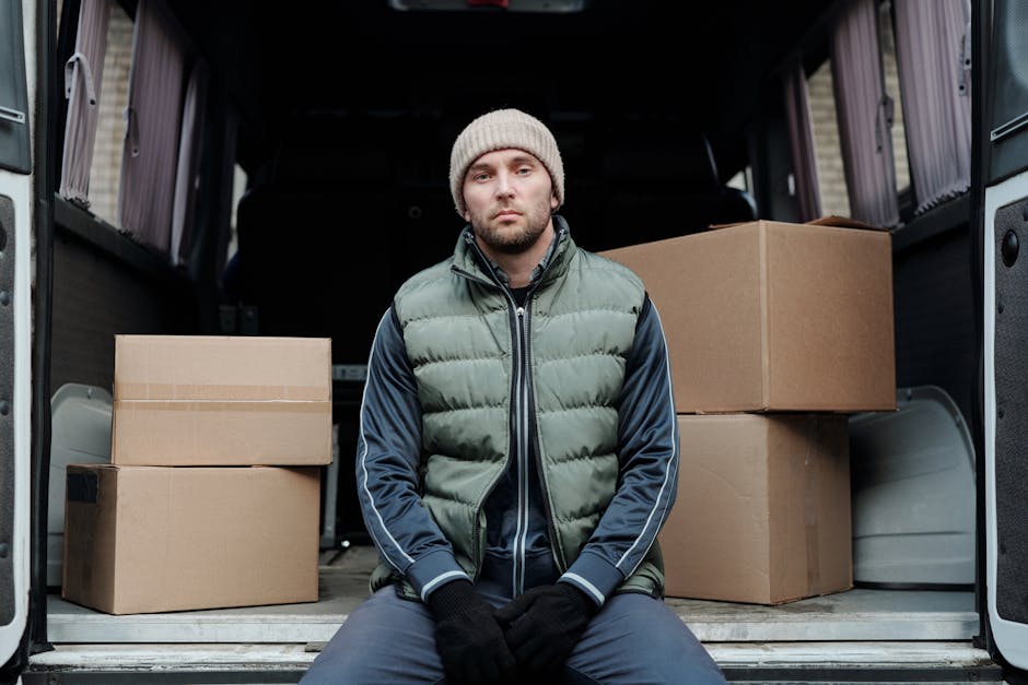 A male removal worker, dressed in a white t-shirt and navy blue overalls, is seated at the open side door of a white moving van. He is carefully lifting a medium-sized cardboard box with one hand, while holding a rolled-up blueprint or packing list in the other. Several other cardboard boxes, some sealed and others partially open, are arranged around him on the ground near the van’s pavement. The interior of the van is visible, showing more wrapped items and packing materials, such as plastic wraps and blankets, intended for furniture protection during a house relocation. The environment suggests a residential setting, with natural lighting illuminating the scene, as the worker focuses on the loading process, indicative of packing and moving operations carried out by Harringay Man and Van during an assisted home removal. The image highlights the logistical effort involved in furniture transport and packing for a house move, with local access challenges implied by the tight space near the vehicle.